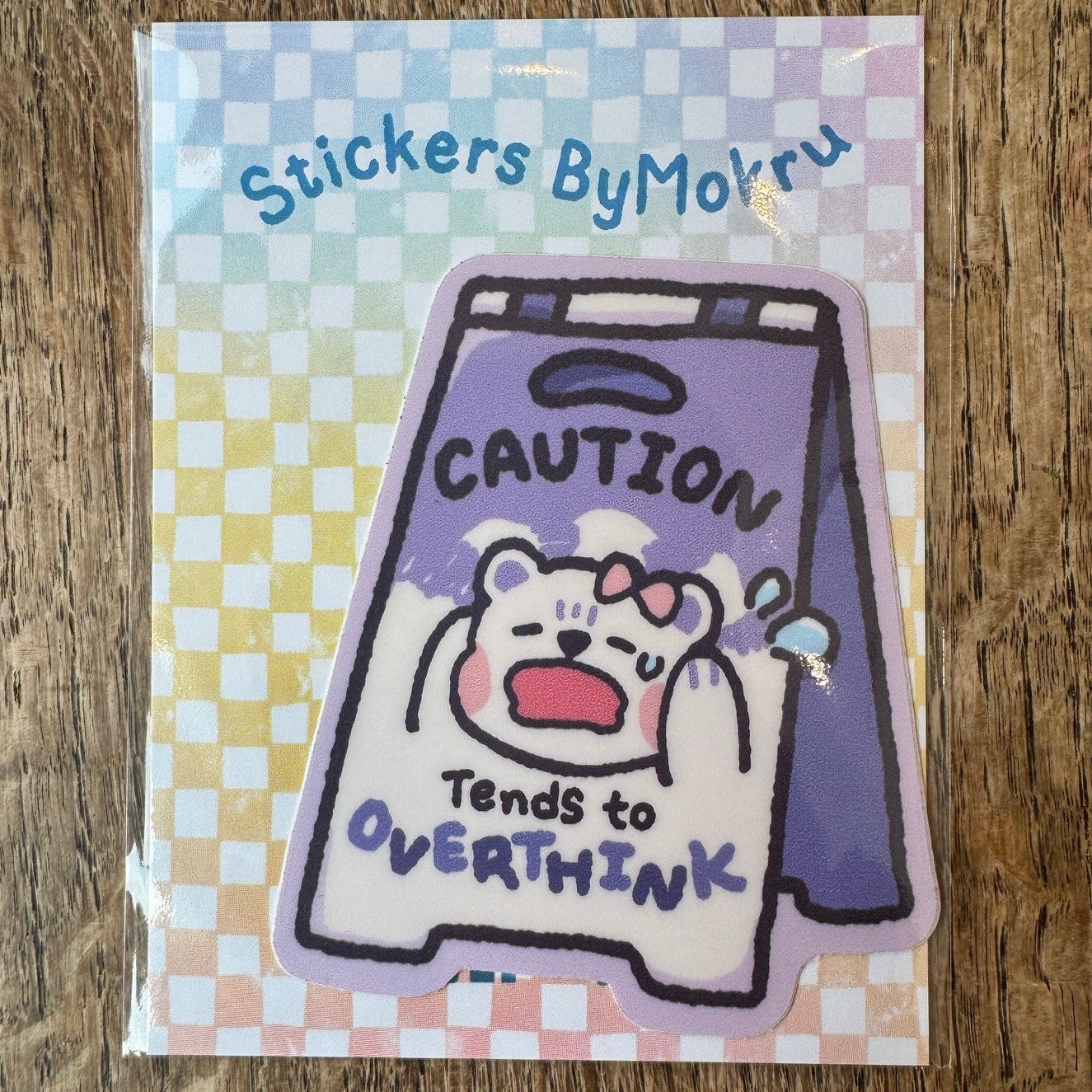 Sticker with a bear illustration and text on a checkered background, placed on a wooden surface.
